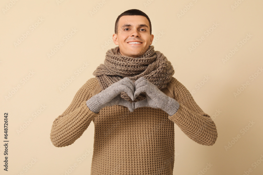 Young man in winter clothes making heart gesture on beige background