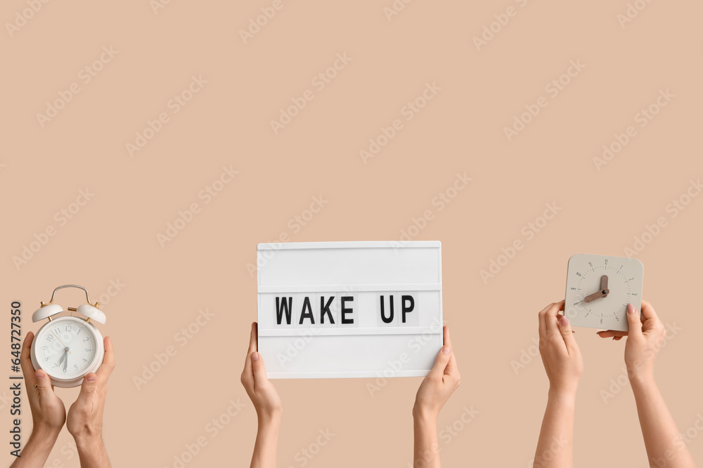 Hands holding alarm clocks and board with text WAKE UP on beige background