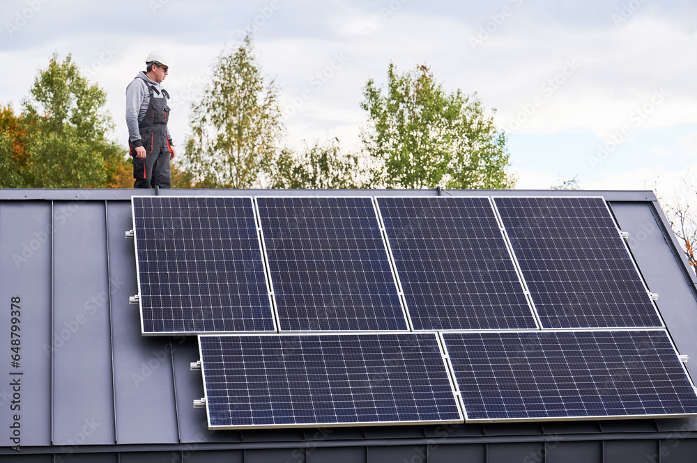 Portrait of man technician installing photovoltaic solar modules on roof of house. Electrician in helmet building solar panel system outdoors. Concept of alternative and renewable energy.