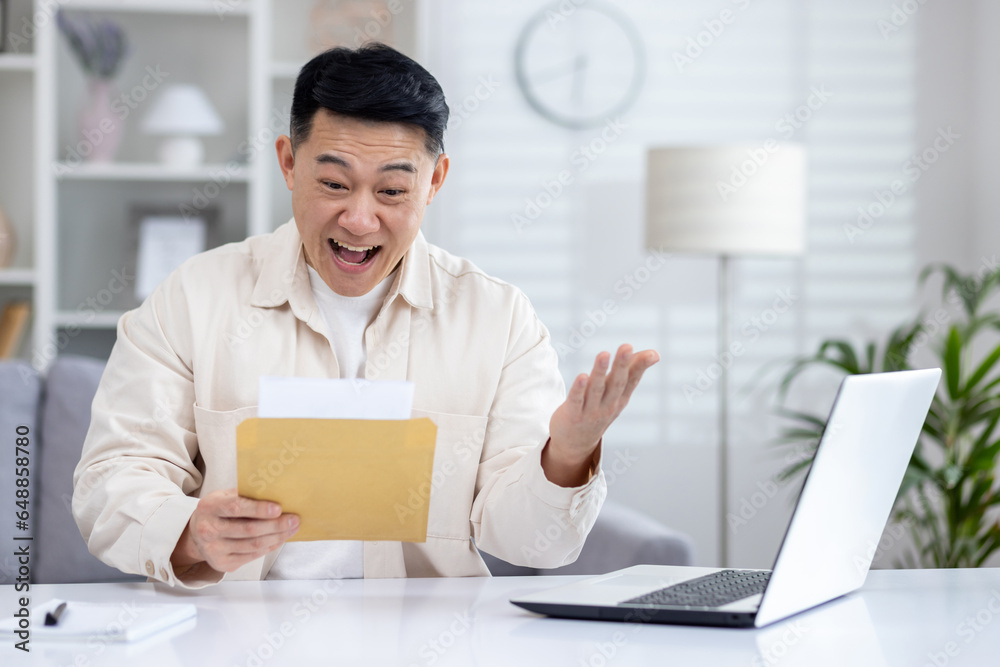 Happy and successful asian received mail notification envelope with good news, man sitting at table at home, working in home office in living room, joyfully celebrating winning news.