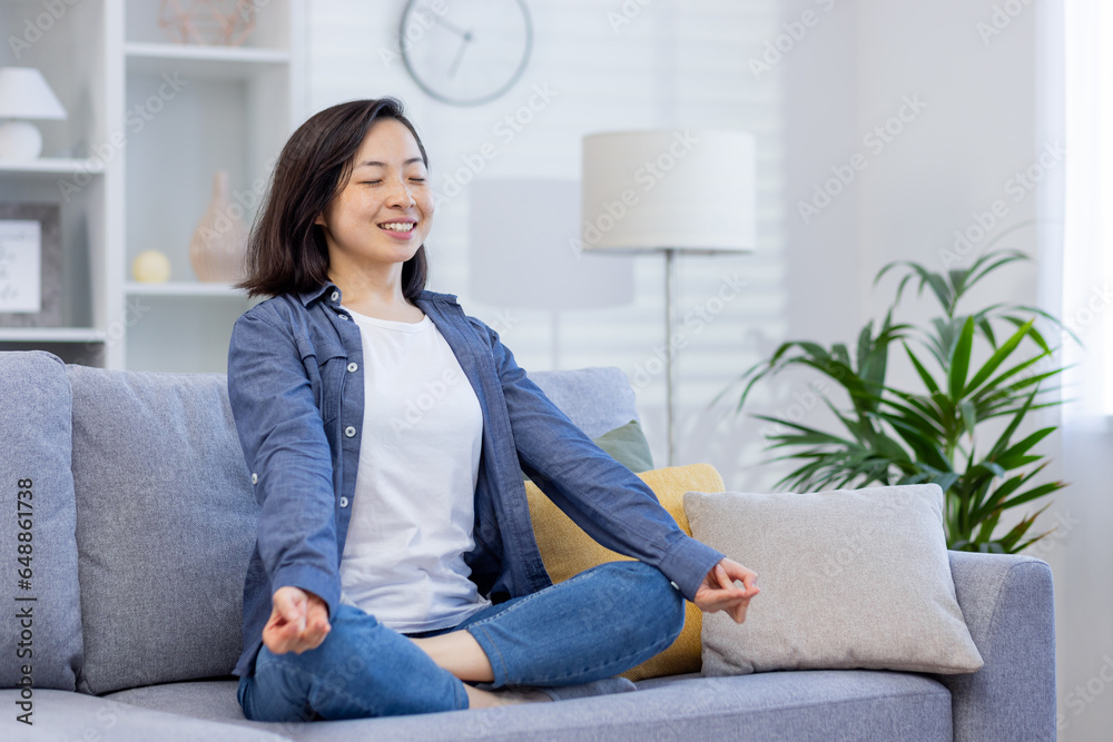 Young smiling asian woman meditating sitting on sofa in living room, woman smiling joyfully with eyes closed, dreaming and thinking about future plans, visualizing achievement.