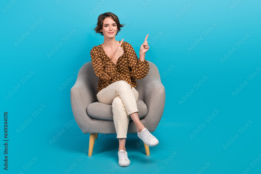 Full body portrait of charming positive lady sit comfort chair indicate fingers empty space isolated on blue color background