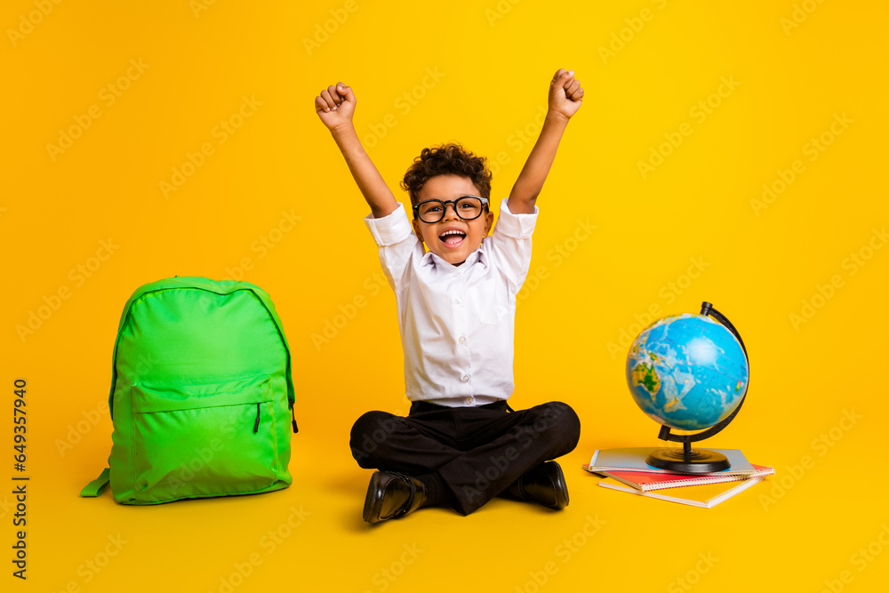 Full body photo of little overjoyed schoolchild sit floor raise fists shout yes world globe rucksack book isolated on yellow color background