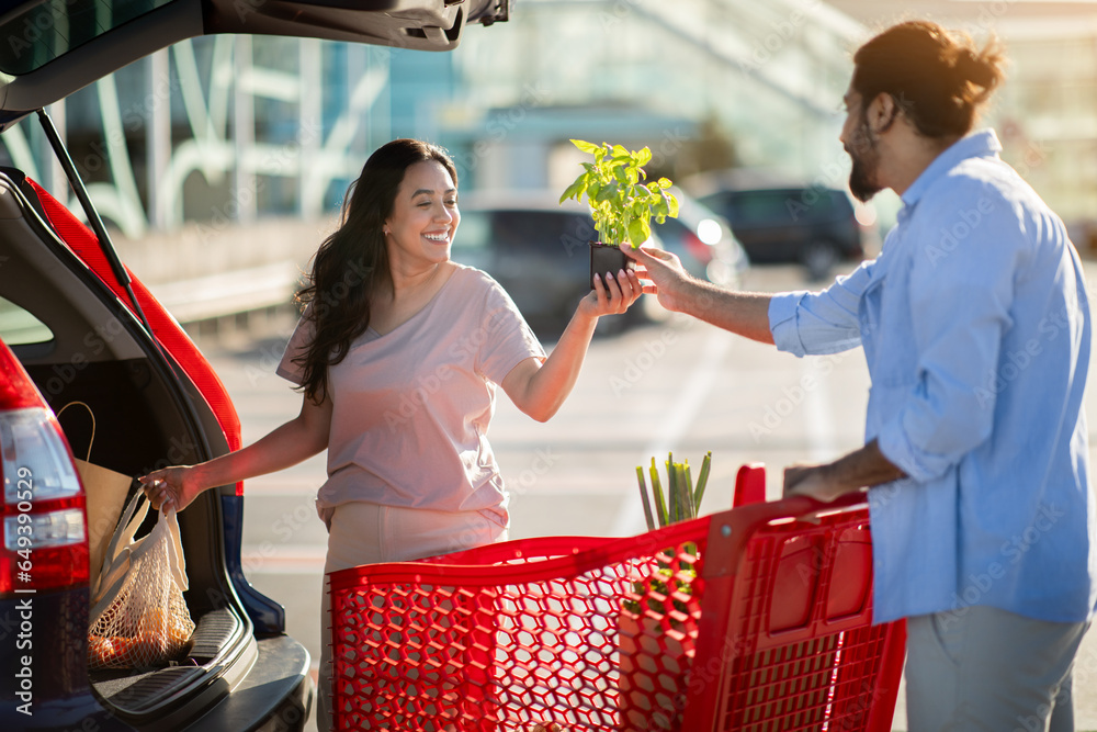 Young happy latin couple with shopping cart full of fresh food, packing products into the car on the outdoor parking