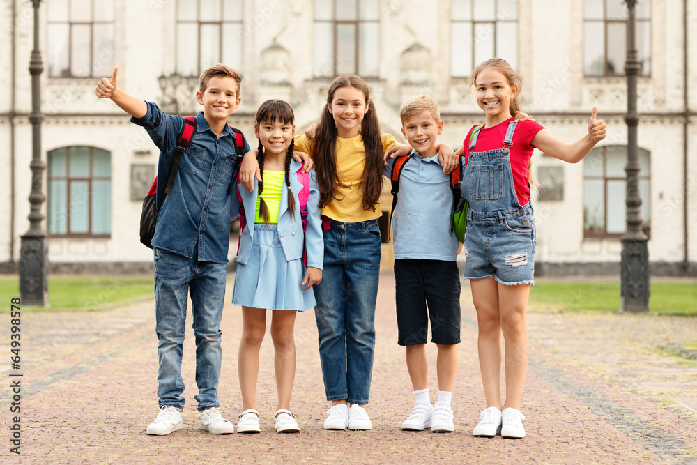 Group of happy multiethnic kids with backpacks posing outdoors, showing thumbs up