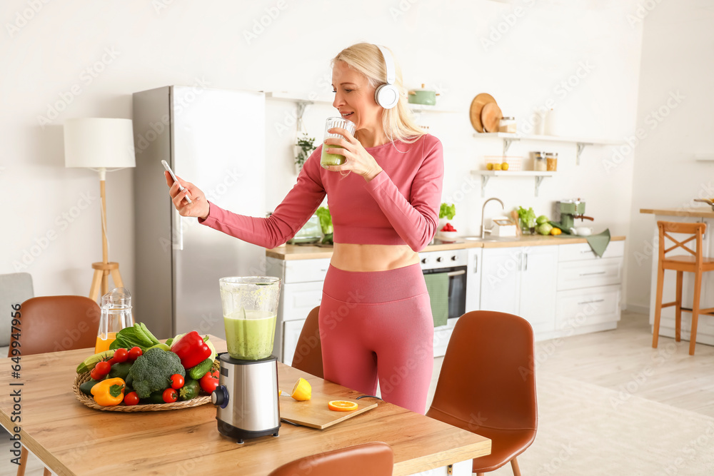 Sporty mature woman with mobile phone and glass of healthy smoothie in kitchen