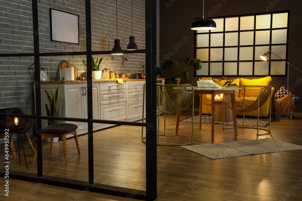 Interior of modern kitchen with glowing lamps, dining table, yellow sofa and white counters at evening