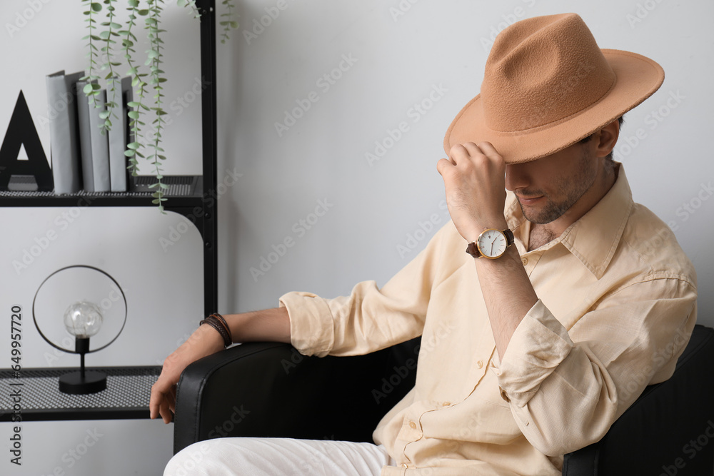 Stylish young man with wristwatch and hat sitting on armchair in room