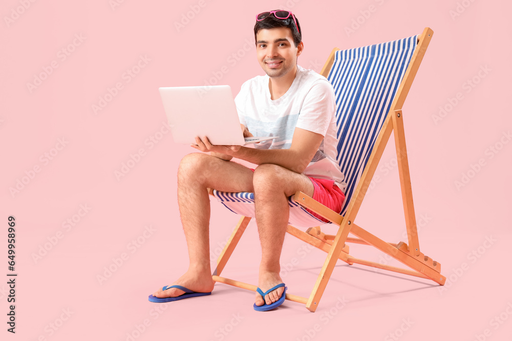 Young man with laptop sitting in deck chair on pink background