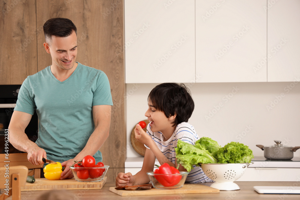 Little boy eating tomato while his father cooking in kitchen