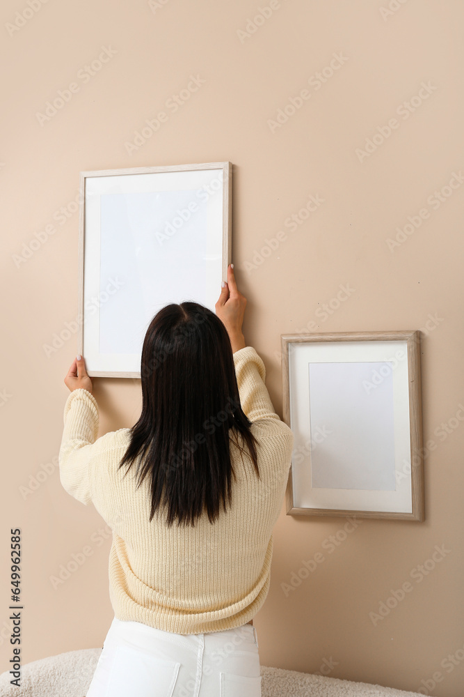 Young woman hanging blank frame on beige wall in living room, back view