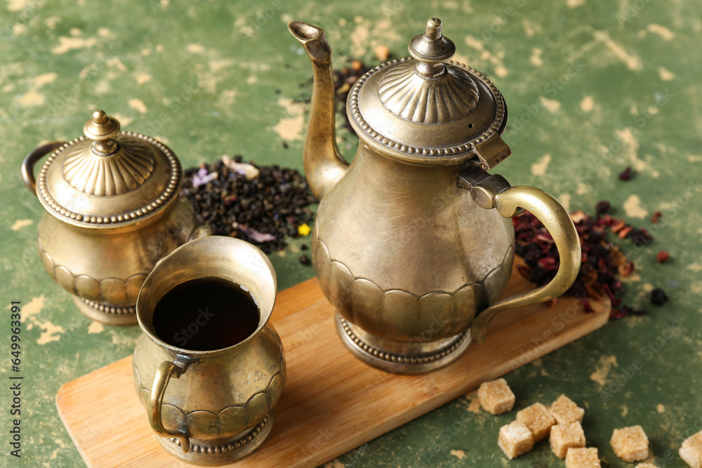 Board with vintage brass tea set, leaves and brown sugar cubes on green table, closeup