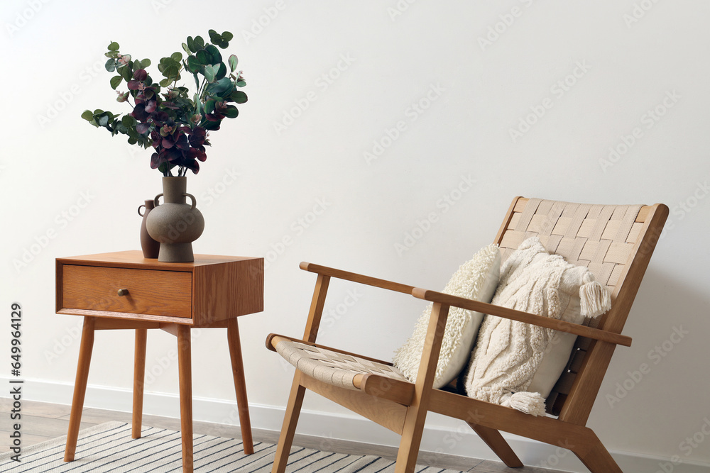 Vase with green eucalyptus branches on end table and comfortable armchair near white wall