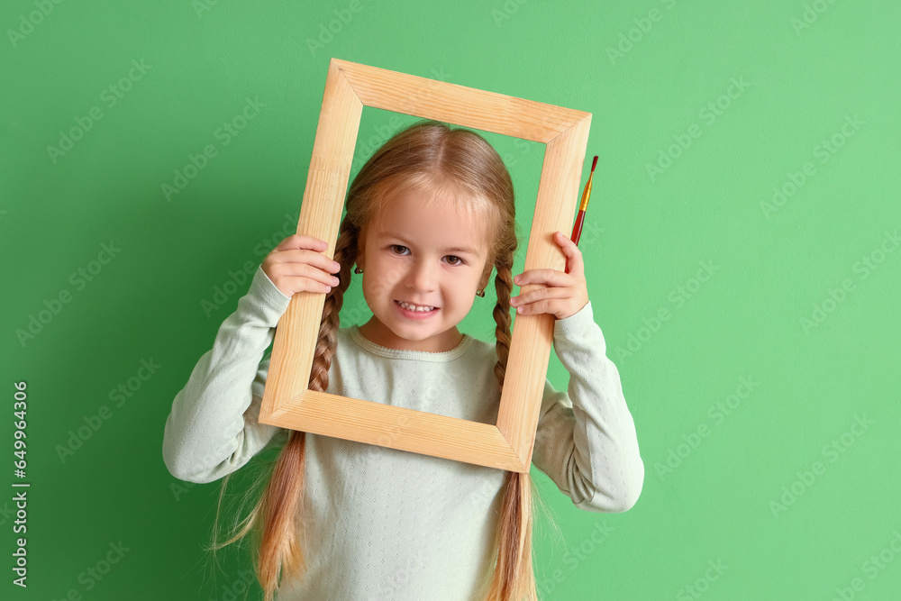 Cute little girl with paint brush and frame on green background