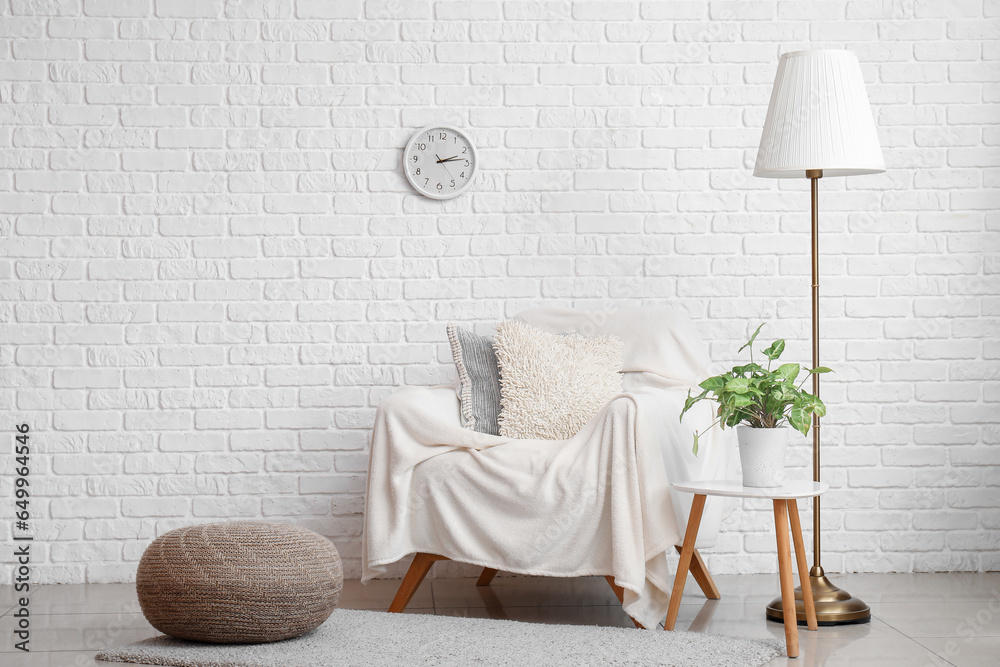 Armchair with blanket, standard lamp and houseplant near white brick wall