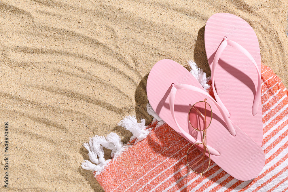 Pair of pink flip flops with sunglasses and blanket on sandy background