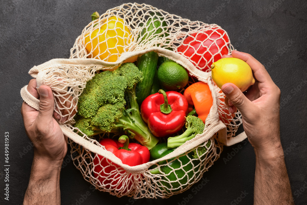 Man putting lemon in mesh bag with different fresh vegetables on black background