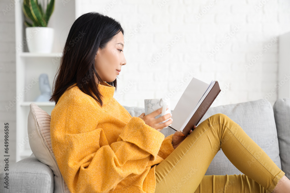 Beautiful young Asian woman with cup of tea sitting on sofa in living room and reading book