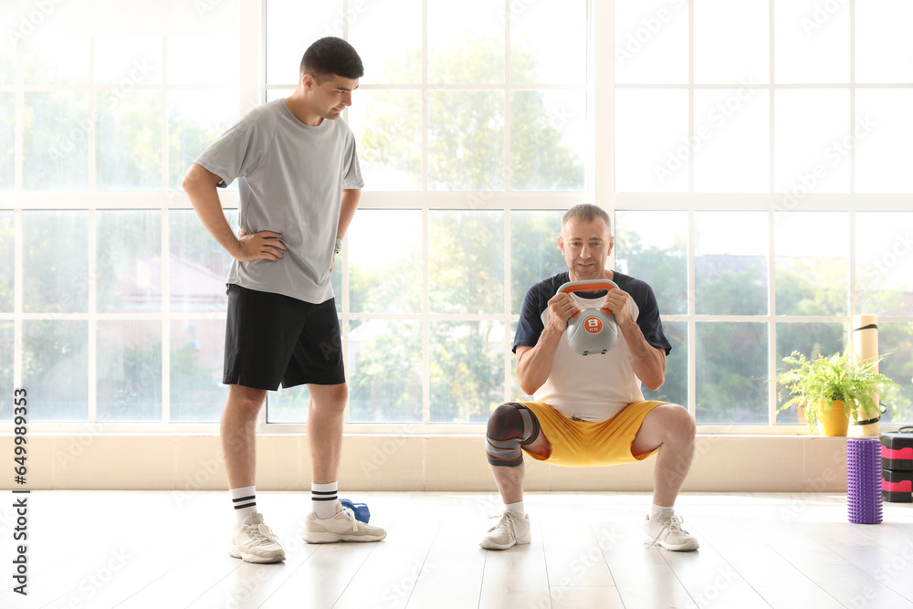 Mature man training with kettlebell and therapist in rehabilitation center