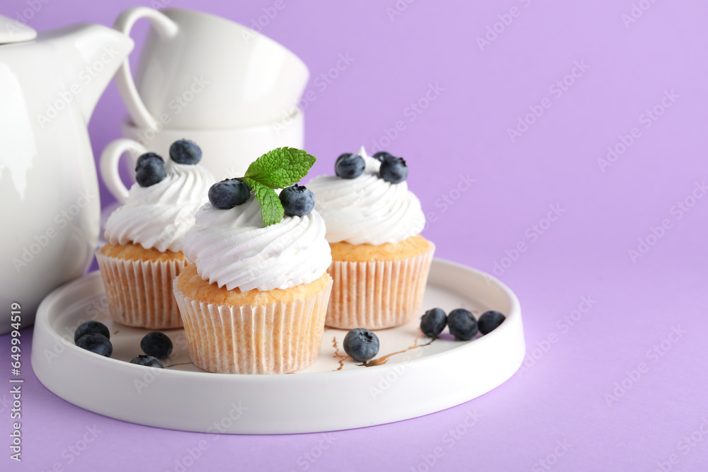 Plate of tasty cupcakes with blueberries on lilac background