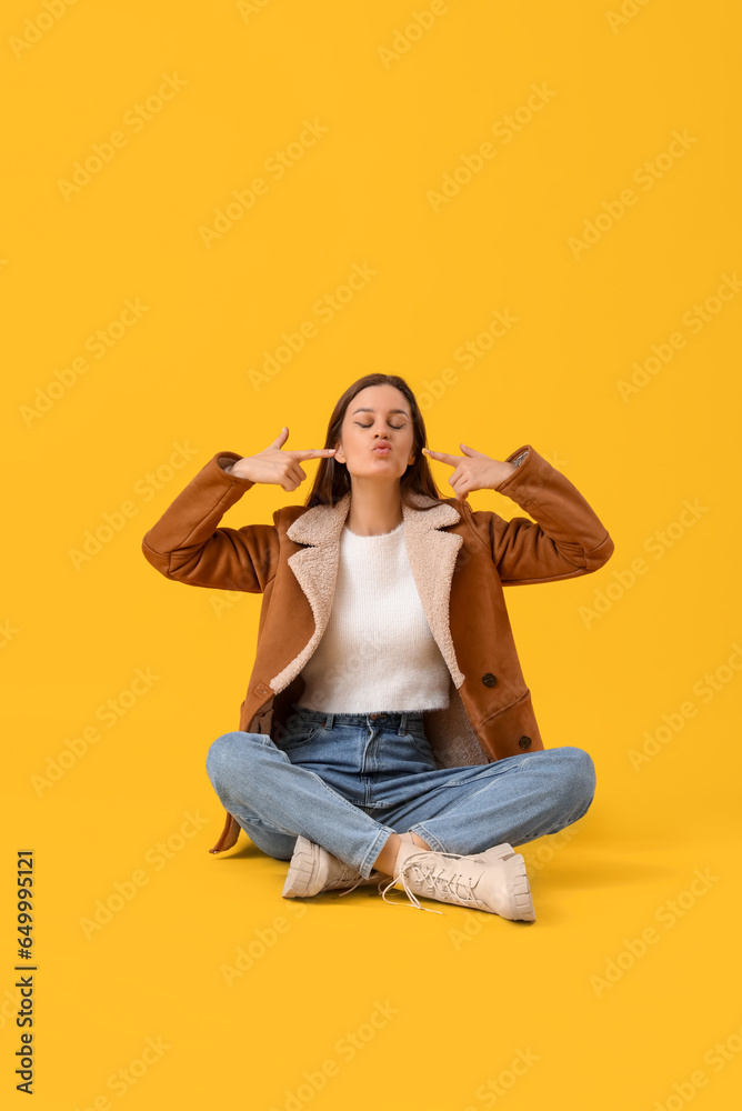 Young woman in winter clothes blowing kiss on yellow background