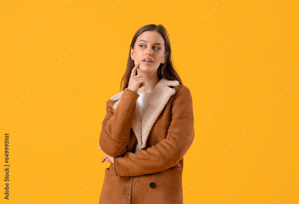 Thoughtful young woman in sheepskin coat on yellow background