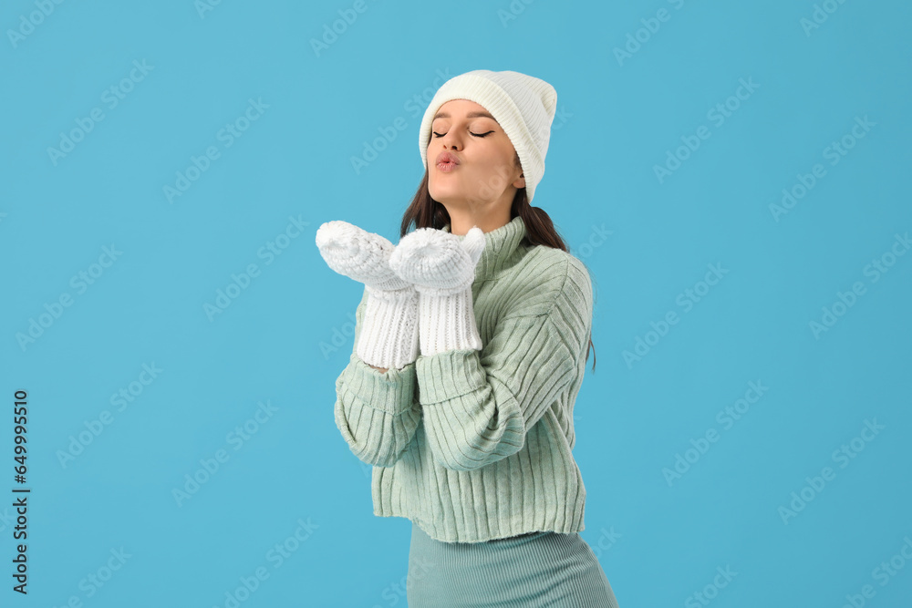 Young woman in winter clothes blowing kiss on blue background