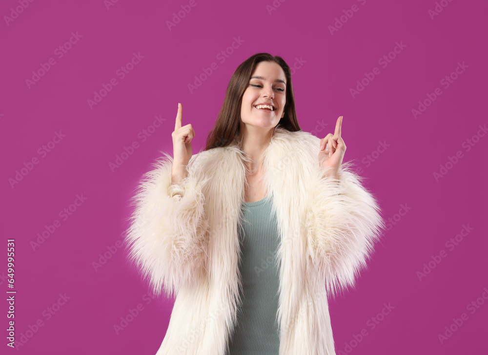 Young woman in fur coat pointing at something on purple background