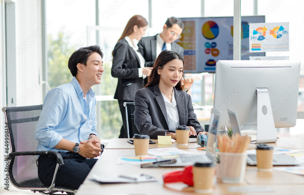 Asian professional successful male businessman mentor supervisor in casual outfit sitting smiling teaching helping female businesswoman intern colleague in formal suit working via computer in office.