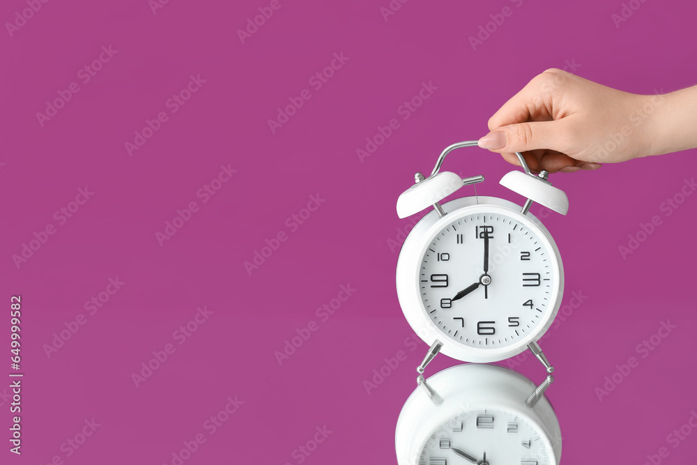 Female hand with alarm clock on table against purple background