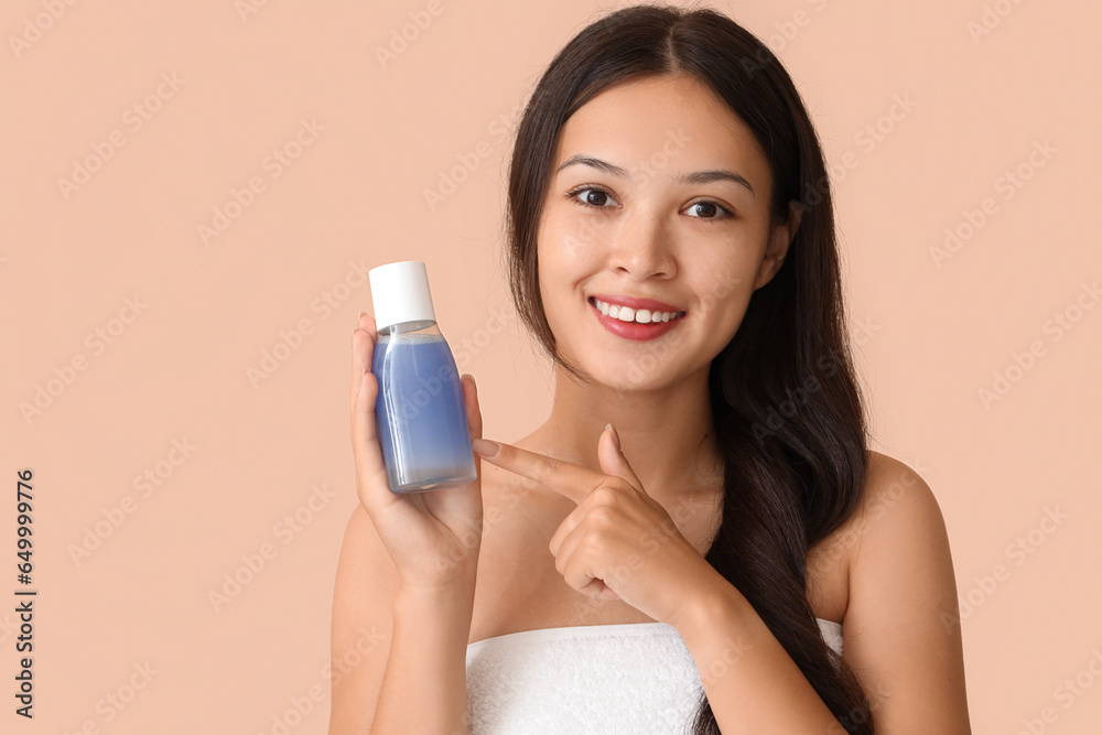 Young Asian woman pointing at bottle of micellar water on beige background, closeup