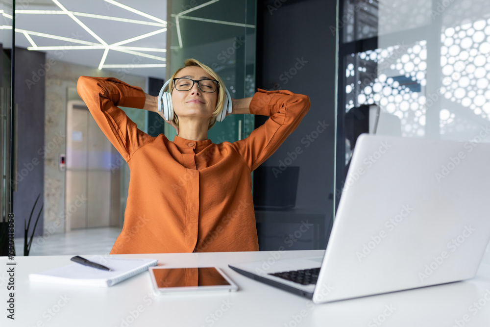 Young beautiful businesswoman relaxing in the office listening to music in headphones, female programmer with her hands behind her head smiling with closed eyes, well done worker completing project.