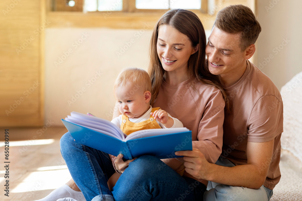 Happy Family Of Three With Cute Infant Son Reading Book Together At Home