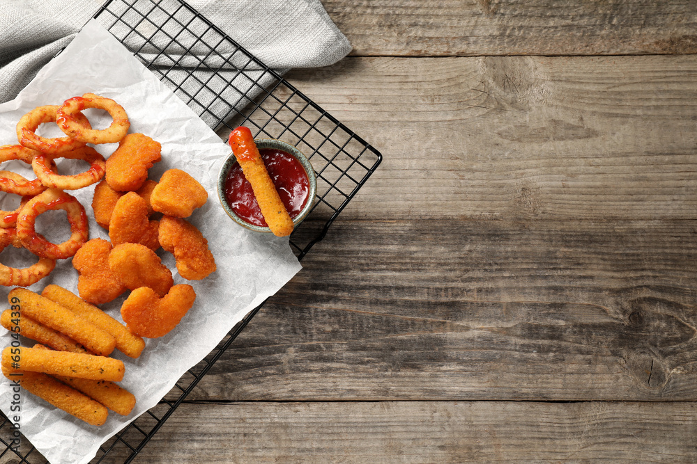 Tasty chicken nuggets, fried onion rings, cheese sticks and ketchup on wooden table, top view. Space for text