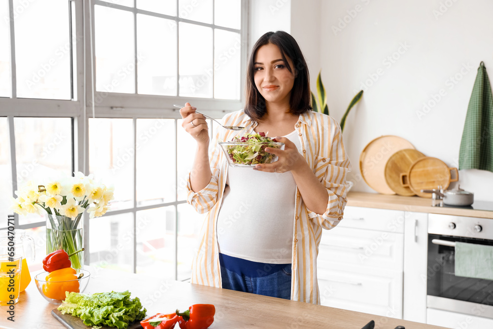 Young pregnant woman eating vegetable salad in kitchen