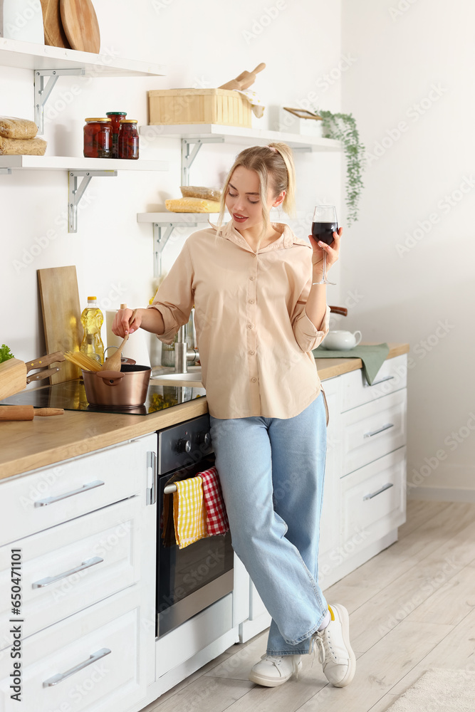 Young woman with glass of wine boiling spaghetti in kitchen