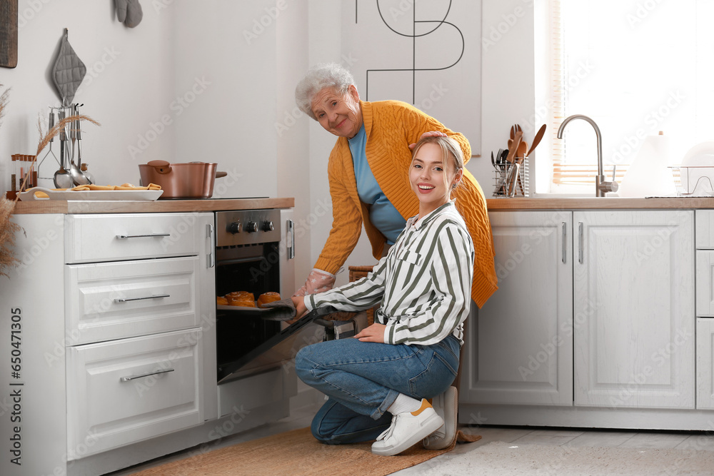 Young woman with her grandmother taking baking tray with buns from oven in kitchen