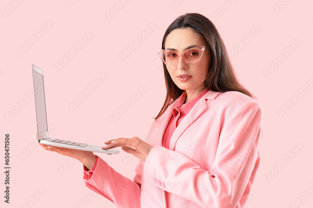 Young woman in stylish suit using laptop on pink background