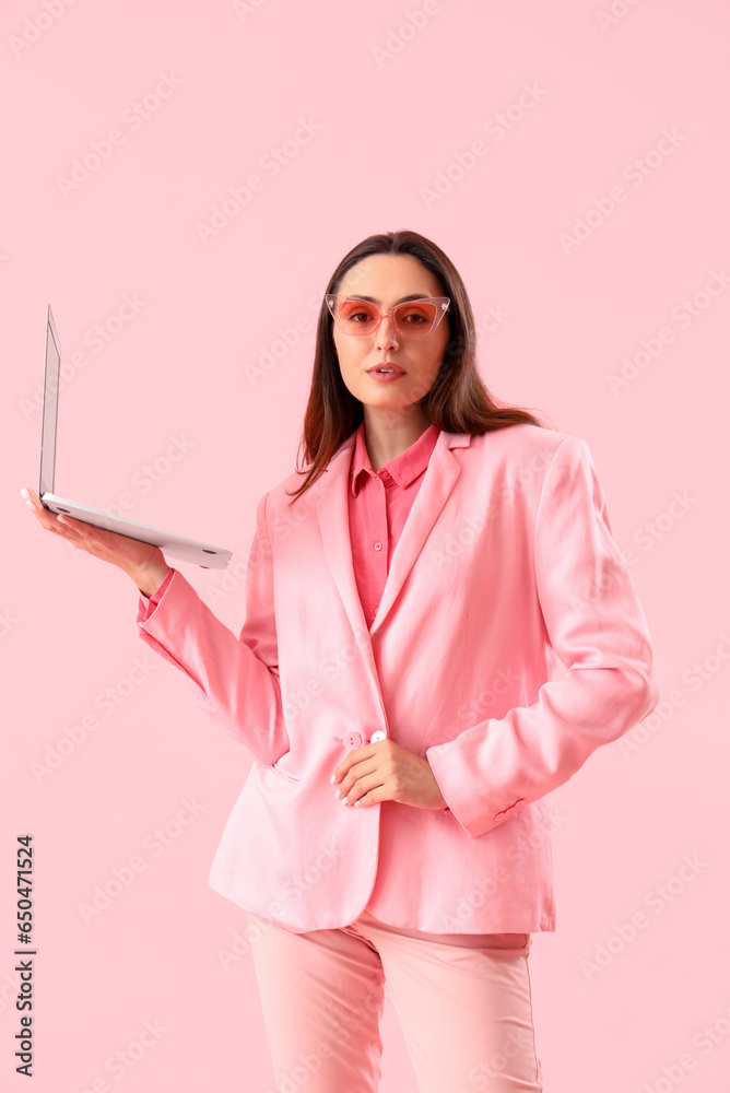 Young woman in stylish suit with laptop on pink background