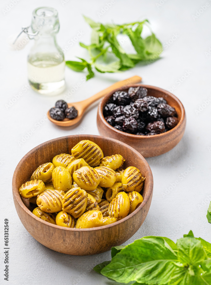 A set of green grill and black dried olives in wooden  bowls on a light background with olive oil and basil close up. The concept of vegetarian healthy snacks