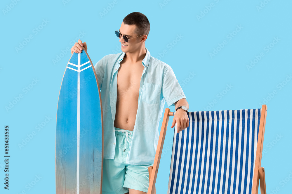 Teenage boy with surfboard and deck chair on blue background