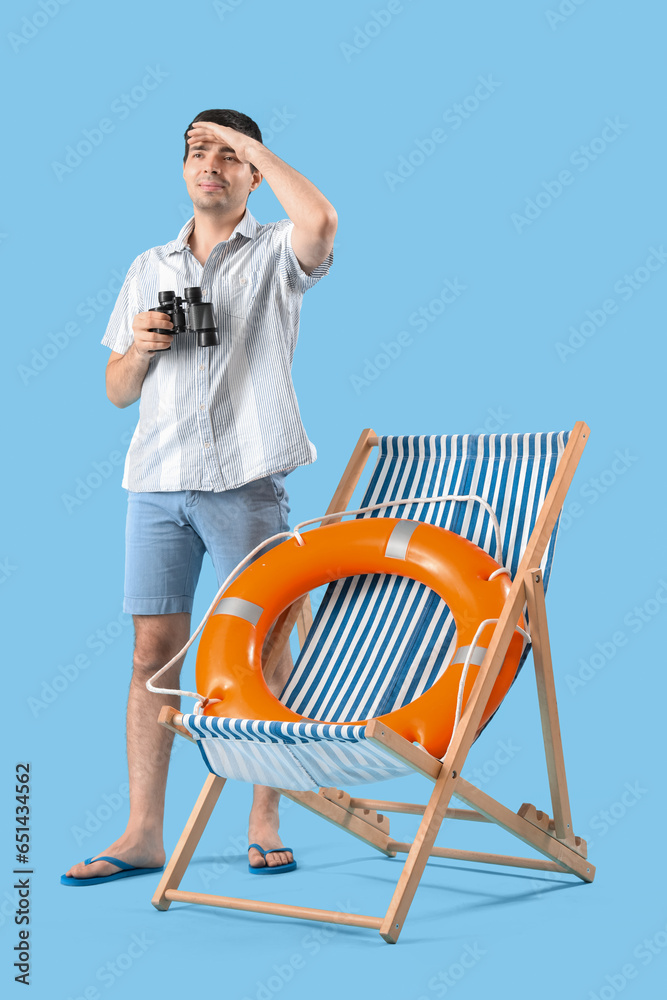 Young man with binoculars, ring buoy and deck chair on blue background