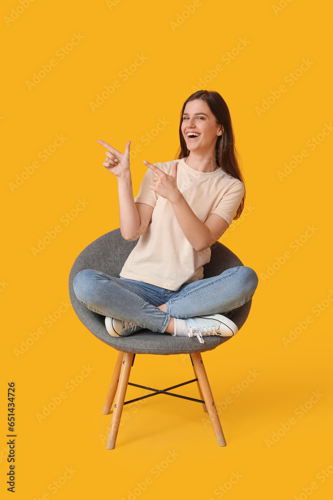 Young woman pointing at something in armchair on yellow background