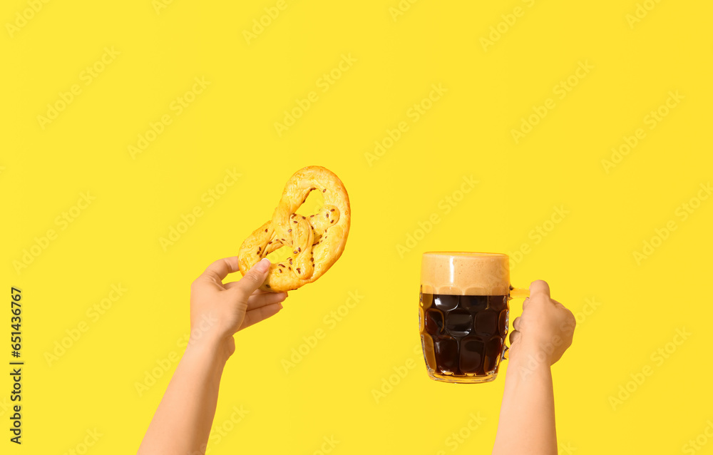 Female hands with mug of cold beer and pretzel on yellow background