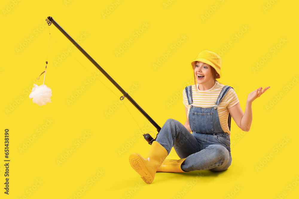 Happy young woman with fishing rod and bath sponge on yellow background