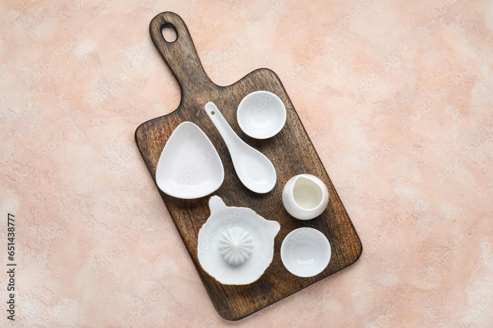 Wooden board with ceramic kitchen utensils on color background