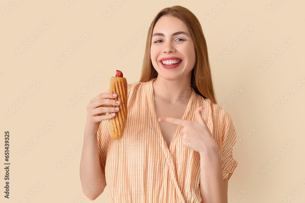 Pretty young woman pointing at tasty french hot dog on beige background