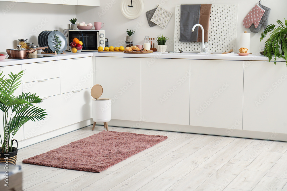 Interior of modern kitchen with stylish rug, trash bin and white counters