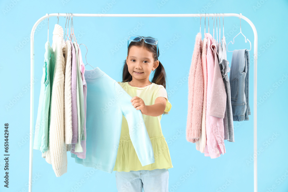 Cute little girl with stylish clothes hanging on rack against light blue background