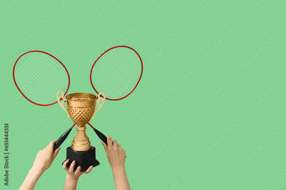 Female hands with gold cup and badminton rackets on green background